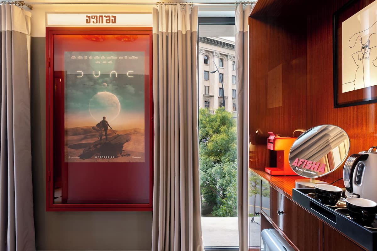 Habitación de hotel con un cartel de 'Dune', vista de un edificio a través de la ventana y una zona de cocina con cafetera y un espejo que muestra 'AFISHA'.