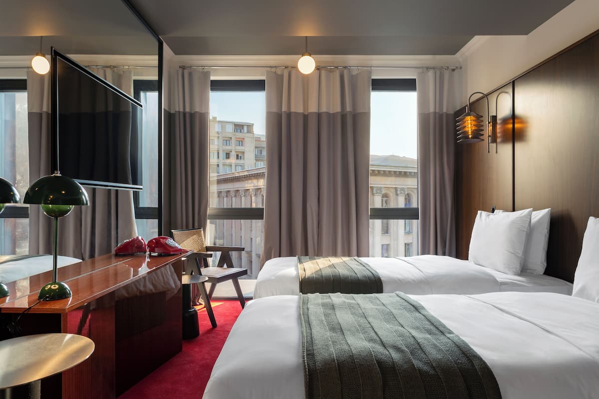 Habitación de hotel con dos camas, un escritorio, una silla y un ventanal grande con cortinas.