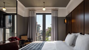 A hotel room with a large bed, a desk with a telephone, a chair, and a view of the city through large windows. - Hotel Afisha (Tbilisi)