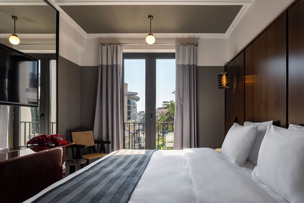 Habitación de hotel con una cama grande, un escritorio con teléfono, una silla y vistas a la ciudad a través de amplios ventanales.