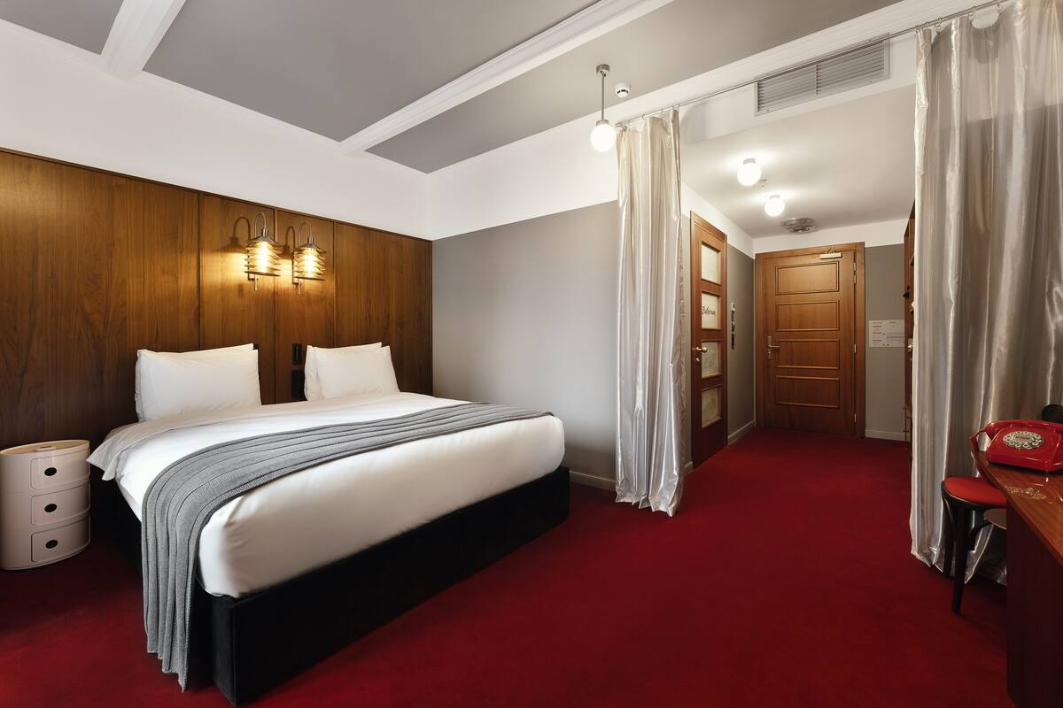 Una habitación de hotel con una cama grande, cabecera de madera, lámparas de noche, alfombra roja, un escritorio con teléfono y una puerta que da a otra habitación.