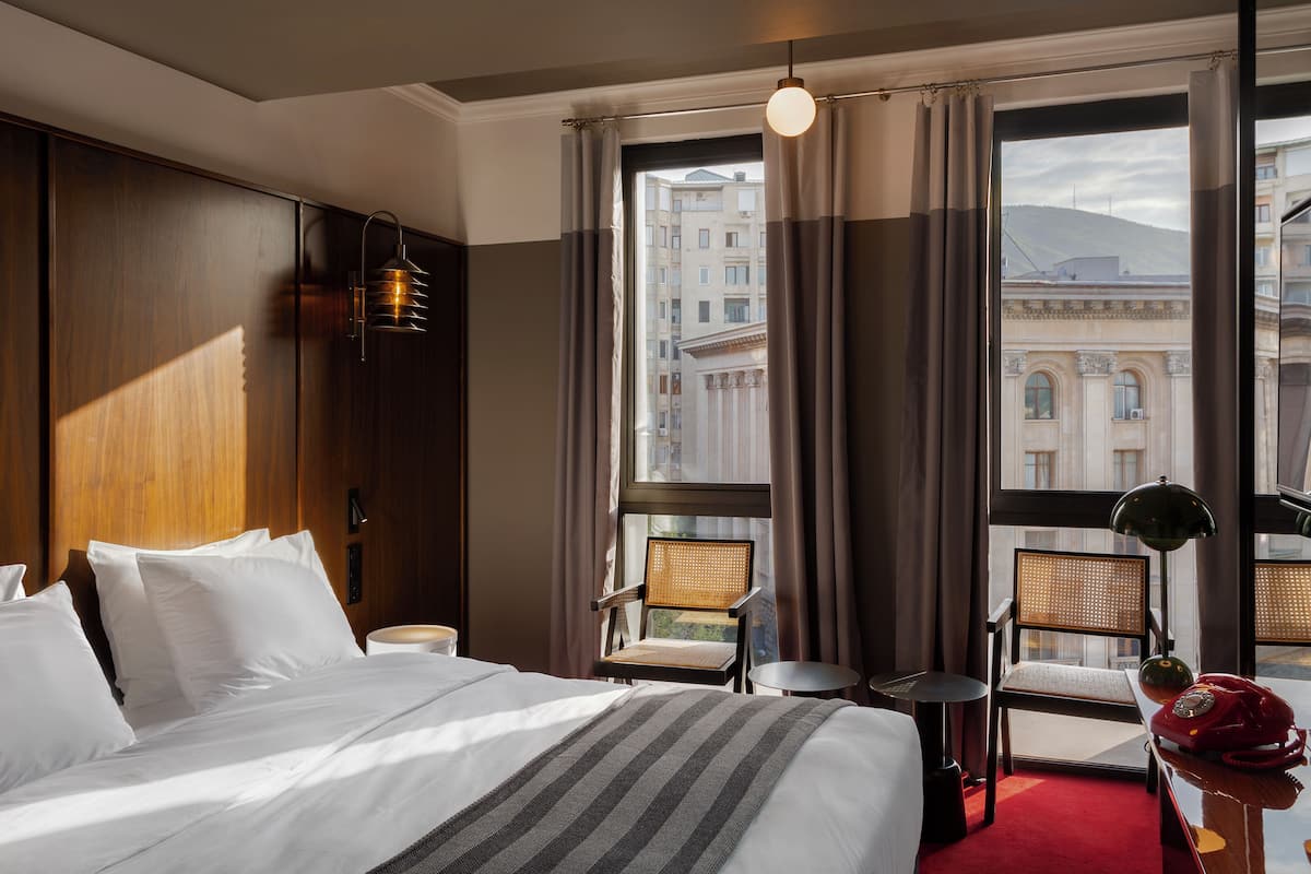 Habitación de hotel con una cama grande, dos sillas, una mesita y vista a un paisaje urbano a través de la ventana.