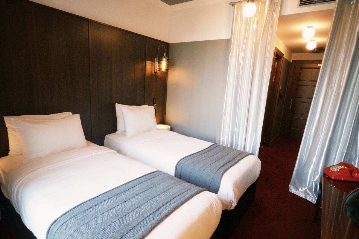 Habitación de hotel con dos camas, cabecero de madera, mesita de noche, teléfono rojo y baño con cortina de ducha.