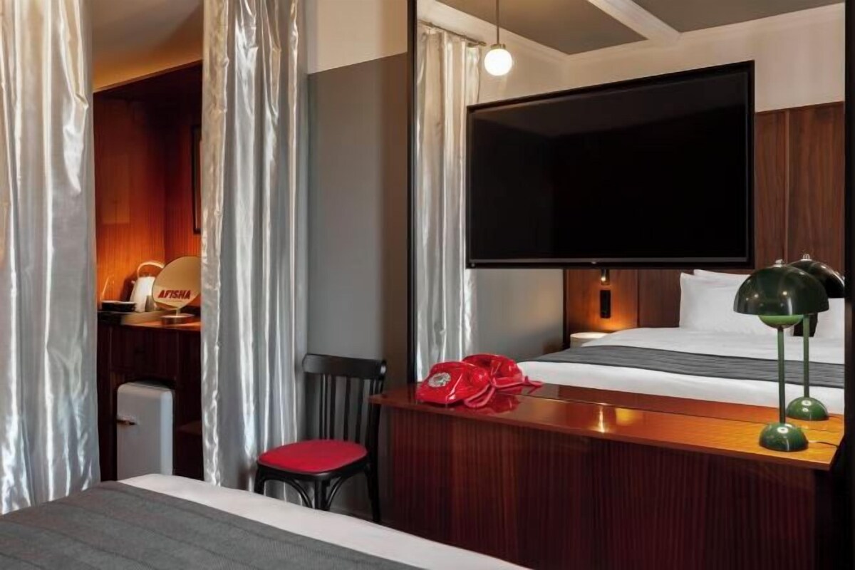 A hotel room with a bed, a desk with a telephone, a television mounted on the wall, and a small desk lamp.