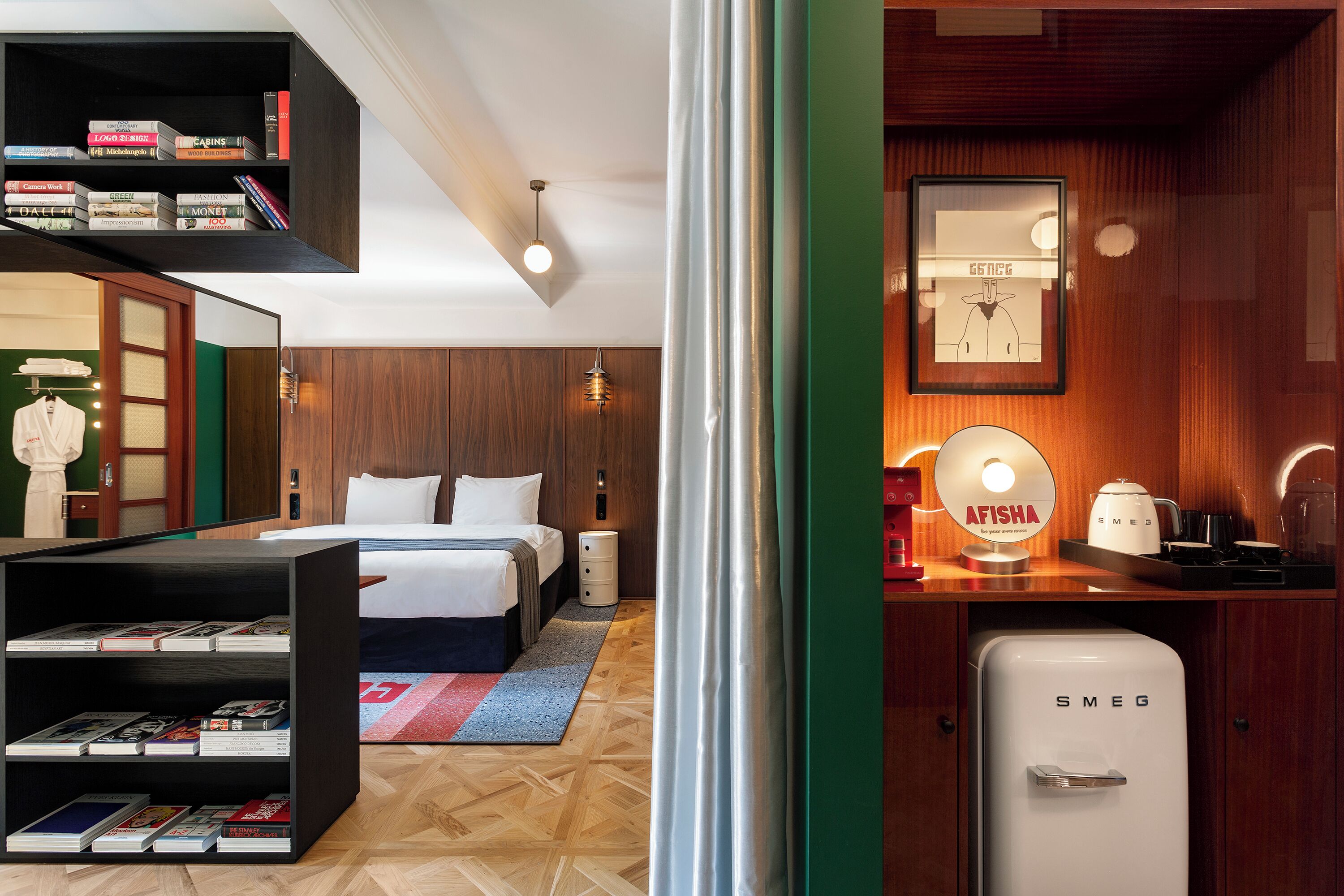 A modern hotel room with a neatly made bed, a bookshelf, a compact kitchenette, and a mirror with a framed picture.