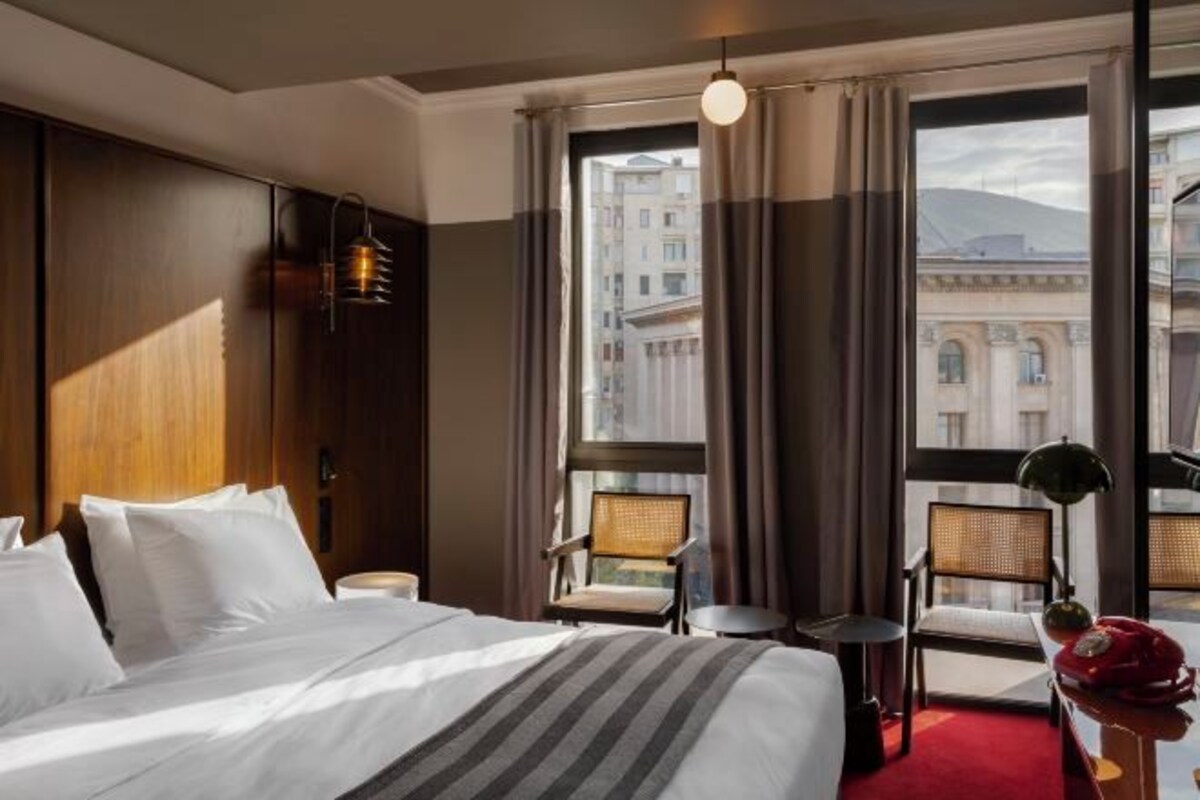 Hotellihuone, jossa on suuri sänky, kaksi tuolia, pieni pöytä ja ikkunasta avautuva näkymä rakennuksiin.