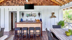 A bar area with a wooden counter, three stools, and a television mounted on the wall. - New Luxury Beachfront Bungalows w/Heated Pool, Pink-Sand Beach (Governors Harbour)