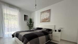 A modern bedroom with a large bed, a bedside table, a nightstand, a floor lamp, and a window with sheer curtains.