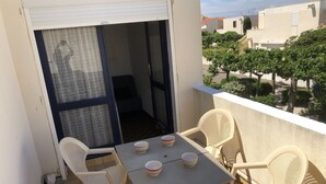 A balcony with a table and four chairs, a sliding glass door, and a view of neighboring buildings and trees.