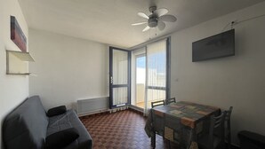 A room with a tiled floor, a grey sofa, a dining table with chairs, a ceiling fan, and a window with curtains.