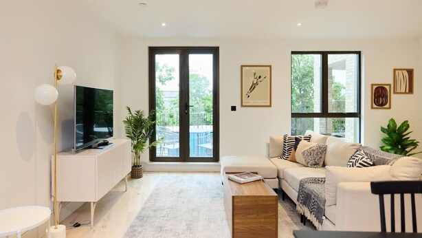 Un salon moderne avec un canapé blanc, une table basse, un téléviseur posé sur un meuble blanc et de grandes fenêtres donnant sur un paysage verdoyant.