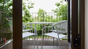 Un balcon avec une table en métal et deux chaises blanches, donnant sur un espace vert arboré.