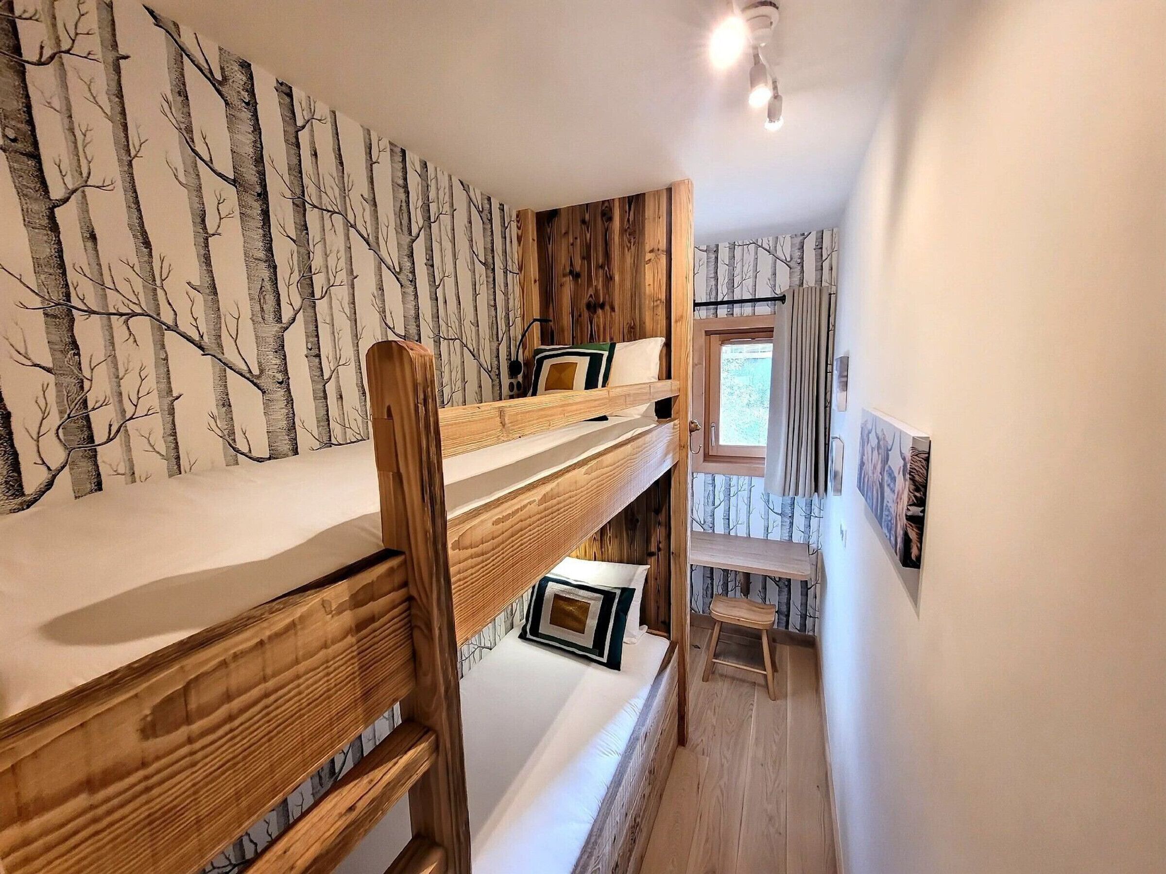 A bunk bed room with a forest wallpaper, a window with curtains, and a small stool.