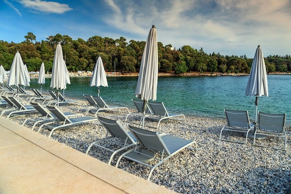 A beachside area with rows of sun loungers and umbrellas, a pebble beach, and a backdrop of trees and water.