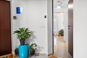 Een gang met een bordje dat een appartement aangeeft, een potplant, een deur, en een kijkje in een woongedeelte met een bank en een kroonluchter.