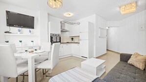 A modern living room with a sofa, a dining table with chairs, a kitchen area with cabinets and a TV mounted on the wall. - Charming White Apartment with Balcony and Parking (Rovinj)
