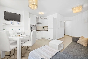 A modern living room with a sofa, a dining table with chairs, a kitchen area with cabinets and a TV mounted on the wall. - Charming White Apartment with Balcony and Parking (Rovinj)