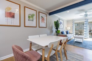 A dining area with a white table and chairs, framed artwork on the wall, and a living room with a sofa and coffee table in the background.