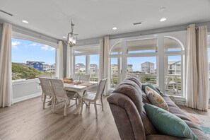 A spacious dining area with a table and chairs, large windows offering a view of the surrounding buildings, and a cozy seating area with a sofa and cushions. - Just Shellaxin #167 - Ocean Views, Private Pool & Hot Tub, Dog Friendly! (North Topsail Beach)