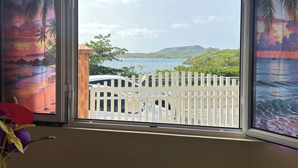 Habitación con vista a un paisaje costero a través de una ventana con paneles decorativos, una valla blanca y una mesa con un objeto rojo.