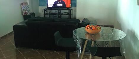 A living room with a TV, a sofa, a glass table with fruit, and a ceiling fan.
