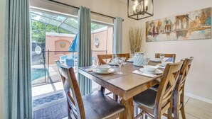 A dining area with a wooden table set for four, surrounded by chairs, with a view of a pool and garden through large windows. - 7 Mi to Disney: Kissimmee Poolside Paradise! (Kissimmee)