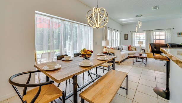 A spacious living area with a wooden dining table, a sofa, and a coffee table. There is a large window providing natural light and a view of the outdoors.