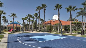 A basketball court with a blue surface, surrounded by palm trees and a building with a red roof.