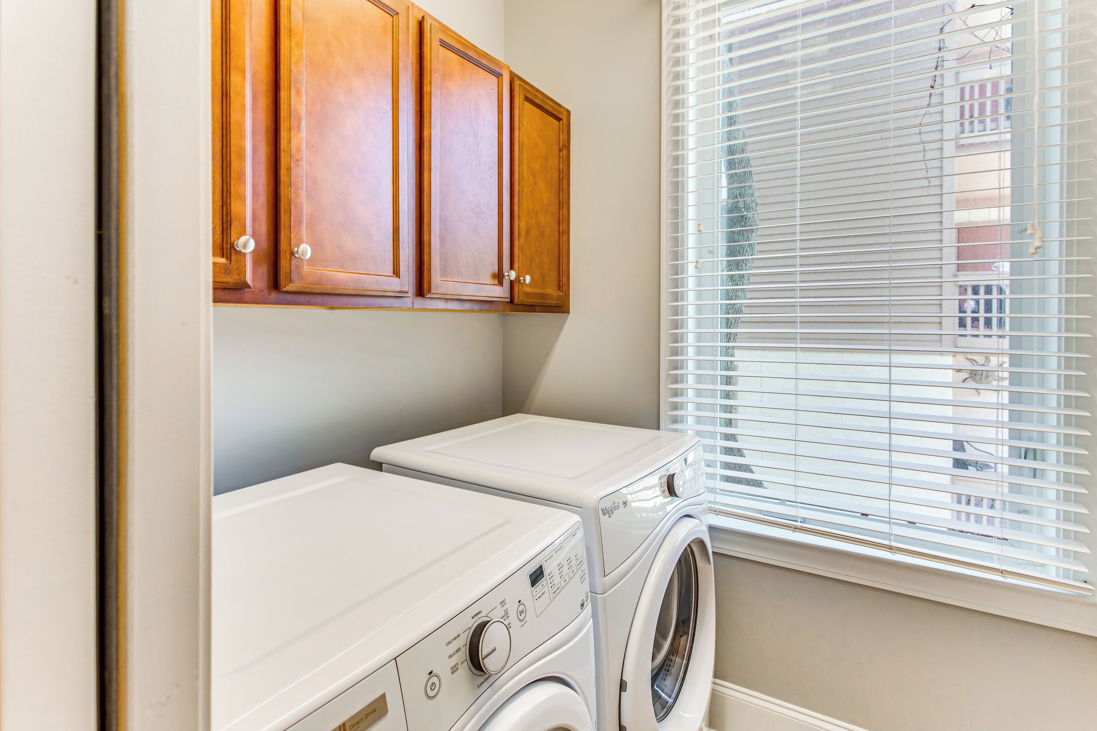 A laundry room with two washing machines, wooden cabinets, and a window with blinds.