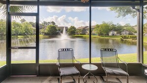 Une terrasse couverte avec deux chaises en métal et une petite table, d’où l’on peut admirer un lac avec une fontaine et des maisons en arrière-plan.