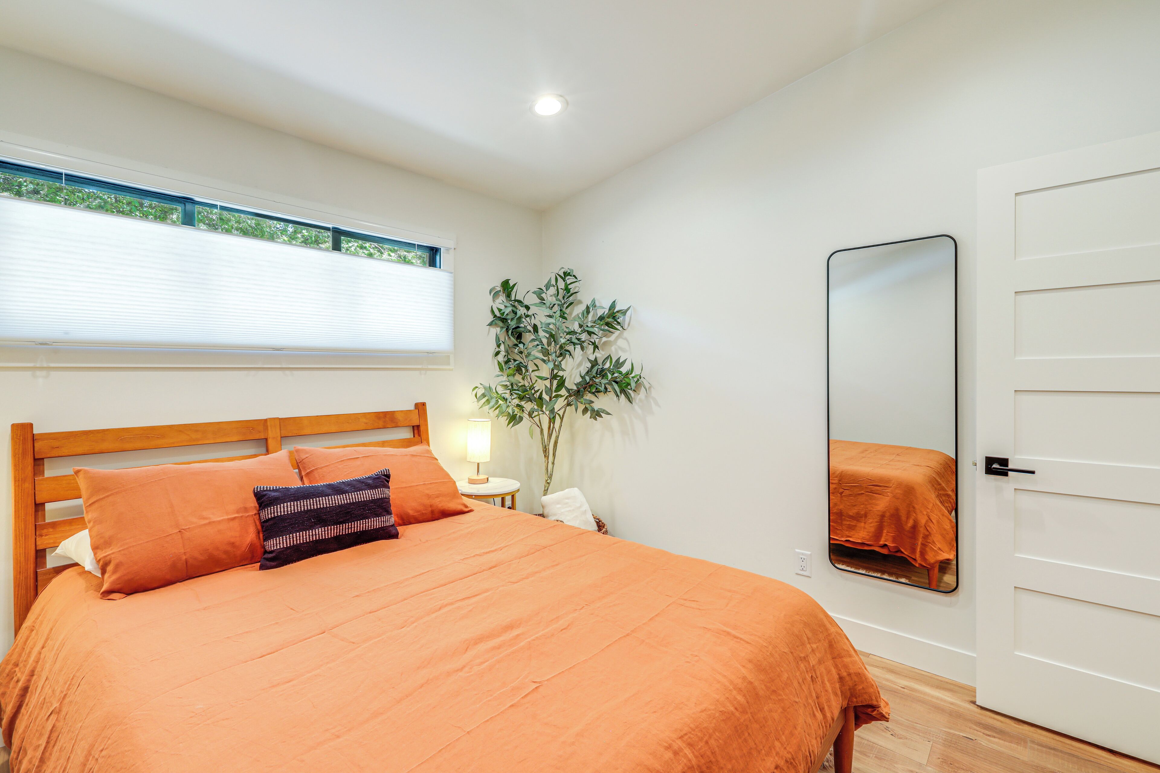 Ein Schlafzimmer mit einem Holzbett, orangefarbener Bettwäsche, einer Nachttischlampe, einem Spiegel und einem Fenster mit Jalousien.