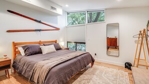 Une chambre à coucher avec un grand lit, une tête de lit en bois, un ventilateur sur pied, une échelle en bois et une fenêtre avec des stores.