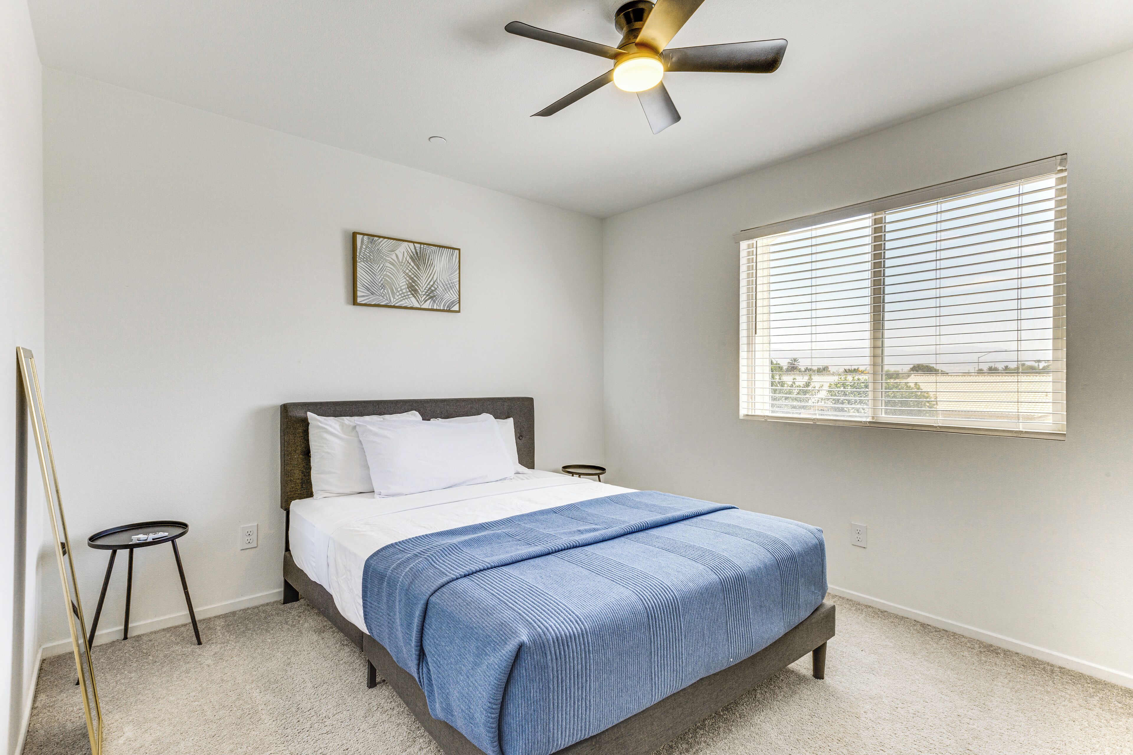 Une chambre à coucher avec un lit, un ventilateur de plafond, une fenêtre avec des stores, une petite table et un tableau accroché au mur.