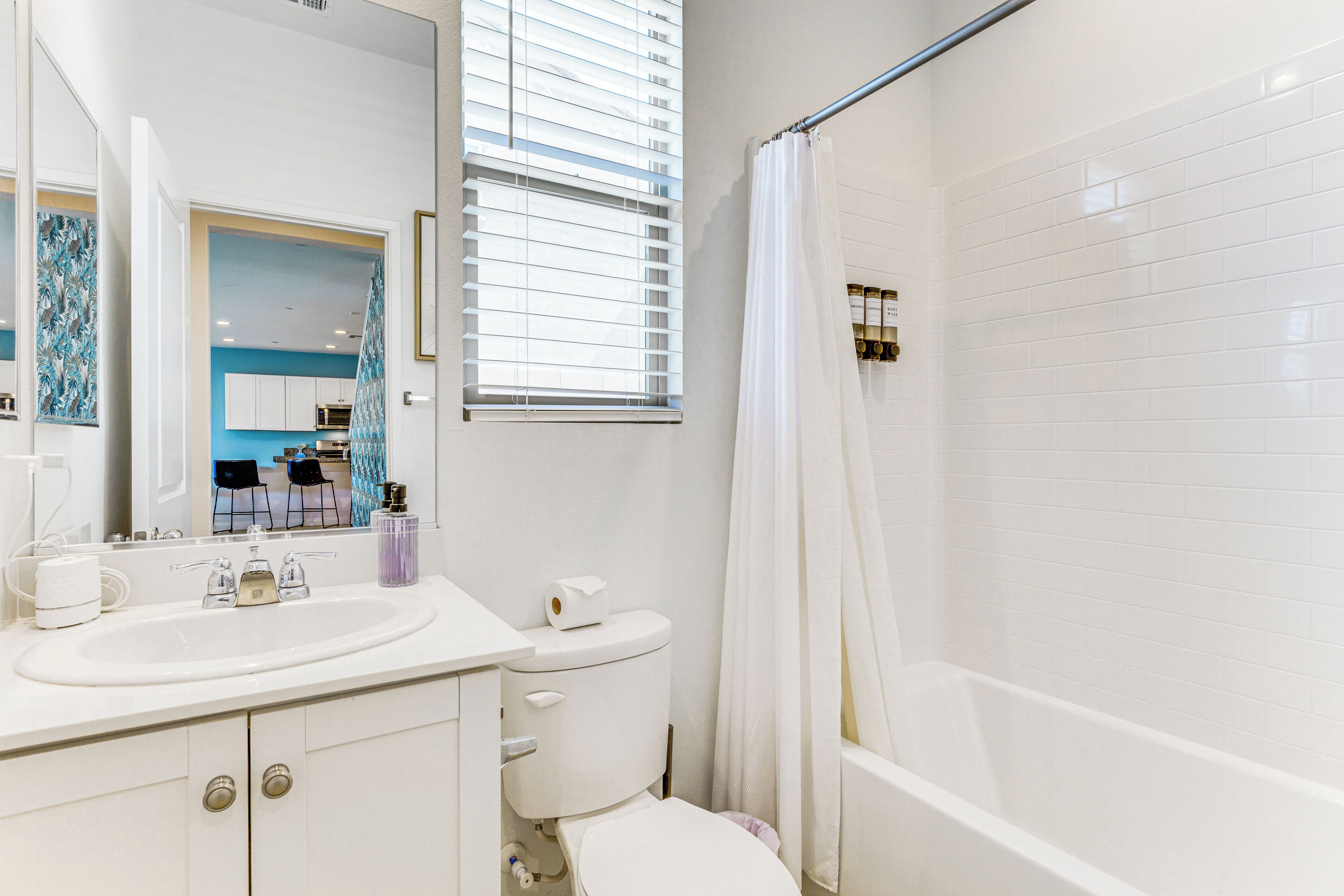 A modern bathroom with a white sink, toilet, and bathtub, a mirror, and a window with blinds.