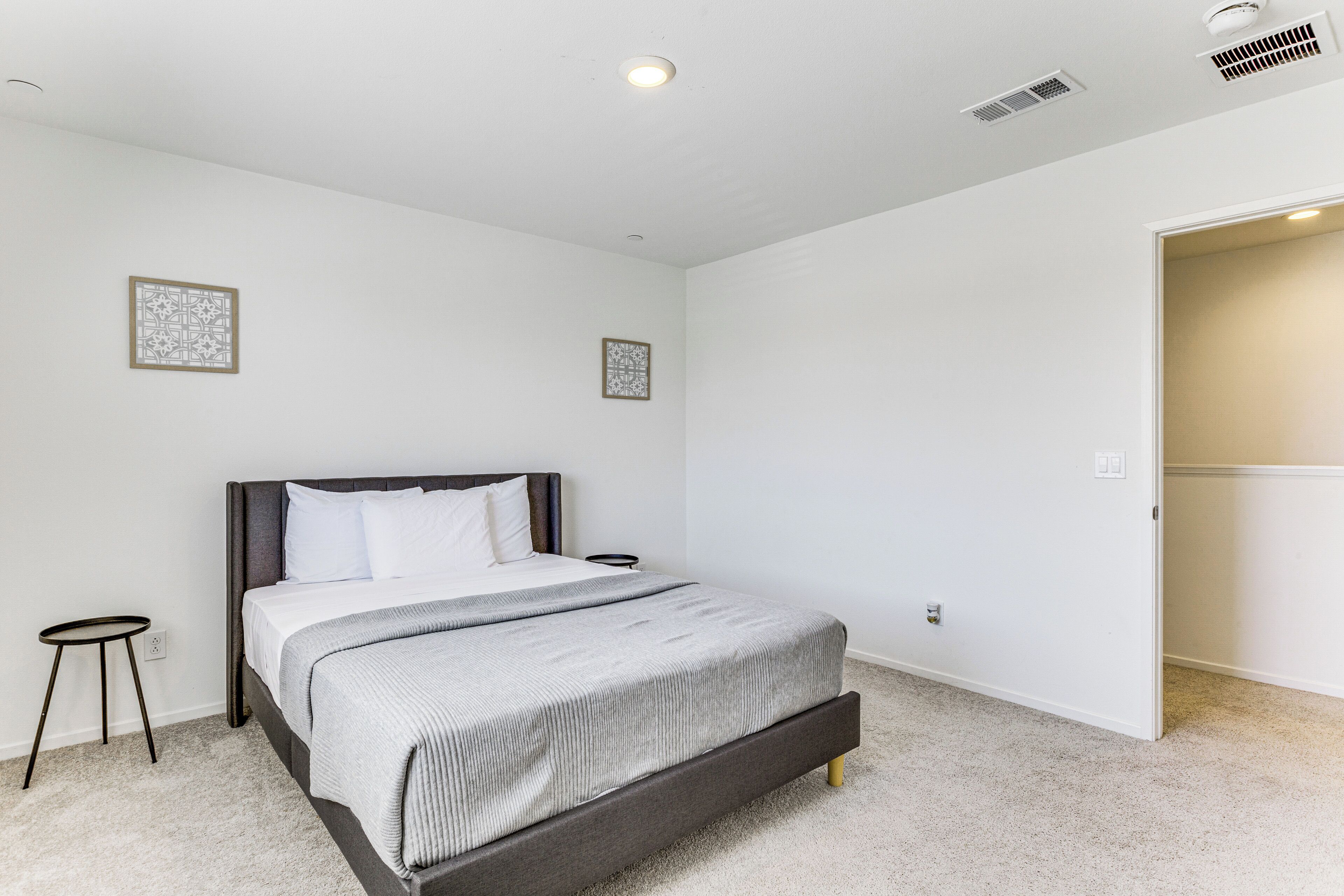 A bedroom with a bed, a small side table, a door leading to another room, and a ceiling vent.
