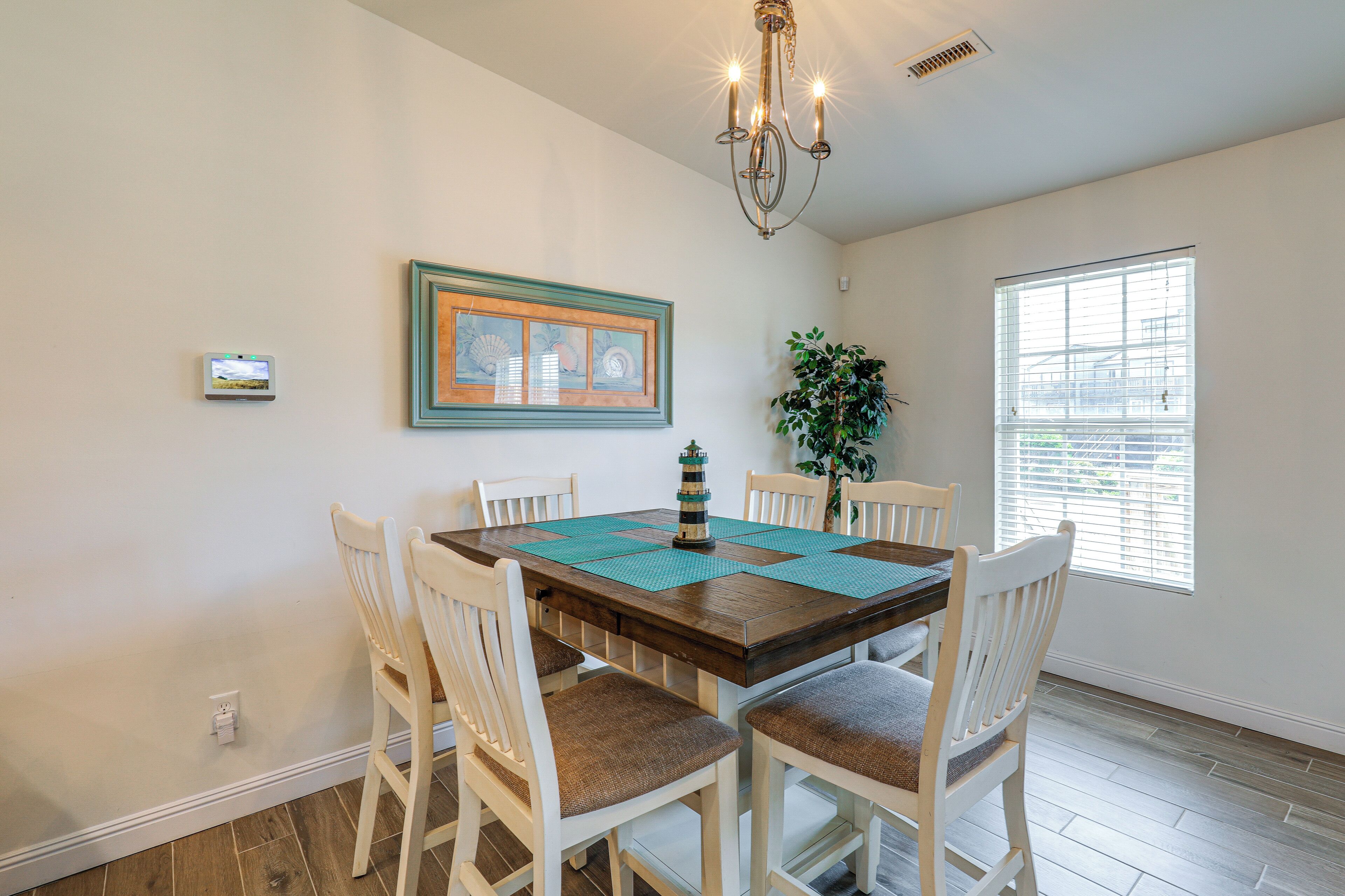 A dining area with a wooden table, white chairs, a chandelier, a framed picture, and a window with blinds.