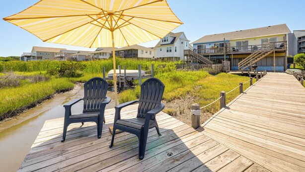 A wooden deck with two chairs and a large striped umbrella, overlooking a grassy area and a water body. - North Topsail Beach Townhome: Walk to the Beach! (Sneads Ferry)