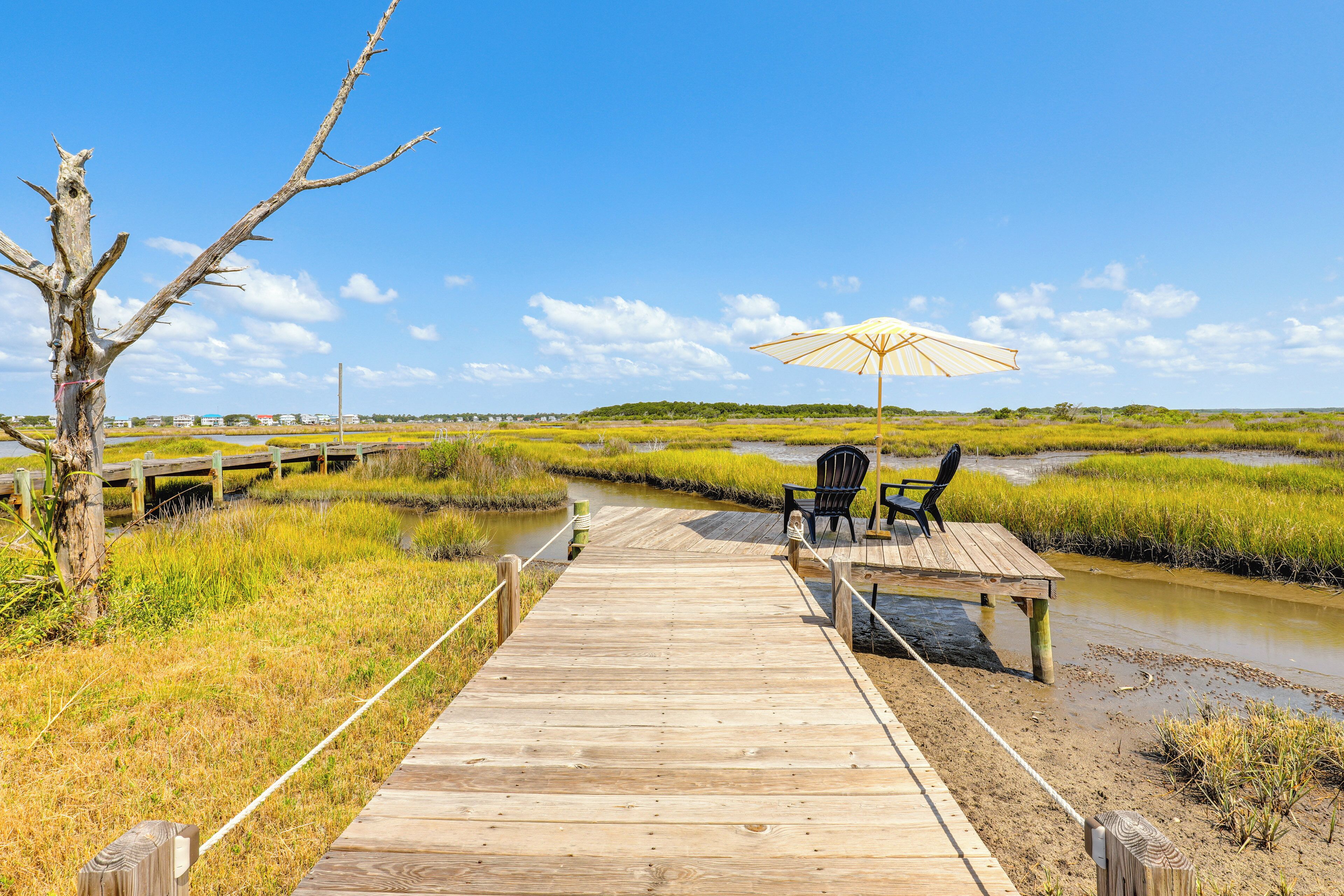 A wooden boardwalk leading to a bench and chair setup with an umbrella, surrounded by marshland and a clear sky.