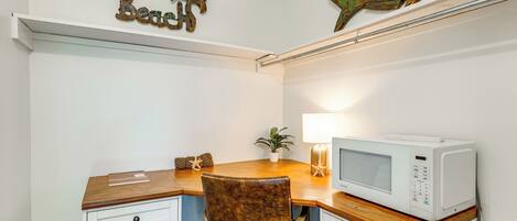 Oficina en esquina con escritorio de madera, una silla blanca, microondas blanco, decoración en la pared y una pequeña planta en maceta.