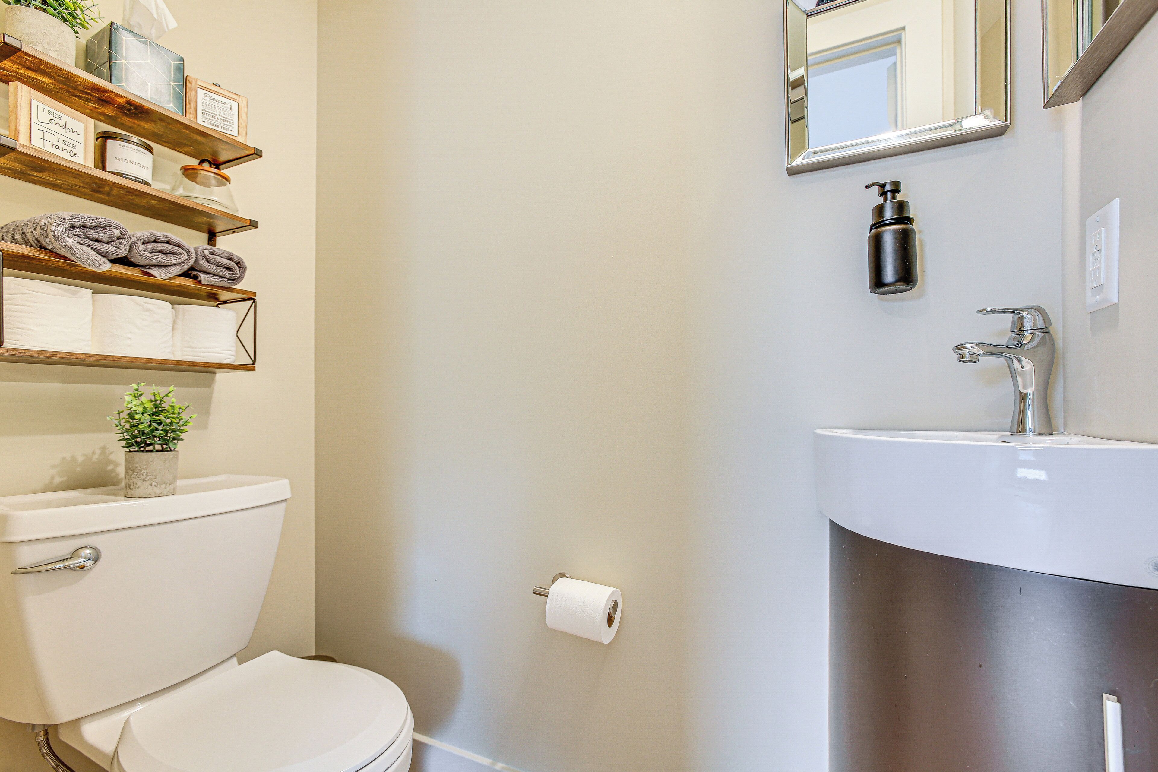 A bathroom with a toilet, a sink, and a mirror. There are shelves with towels and a small plant.