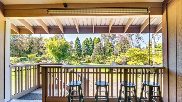 A balcony with a wooden railing, three black barstools, and a view of a garden with various trees and shrubs. - Rainforest Retreat w/ Hot Tub: Mins to Nat'l Park! (Volcano)