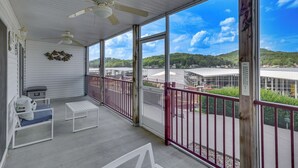 A covered balcony with a view of a lake and hills, white furniture, and a ceiling fan.