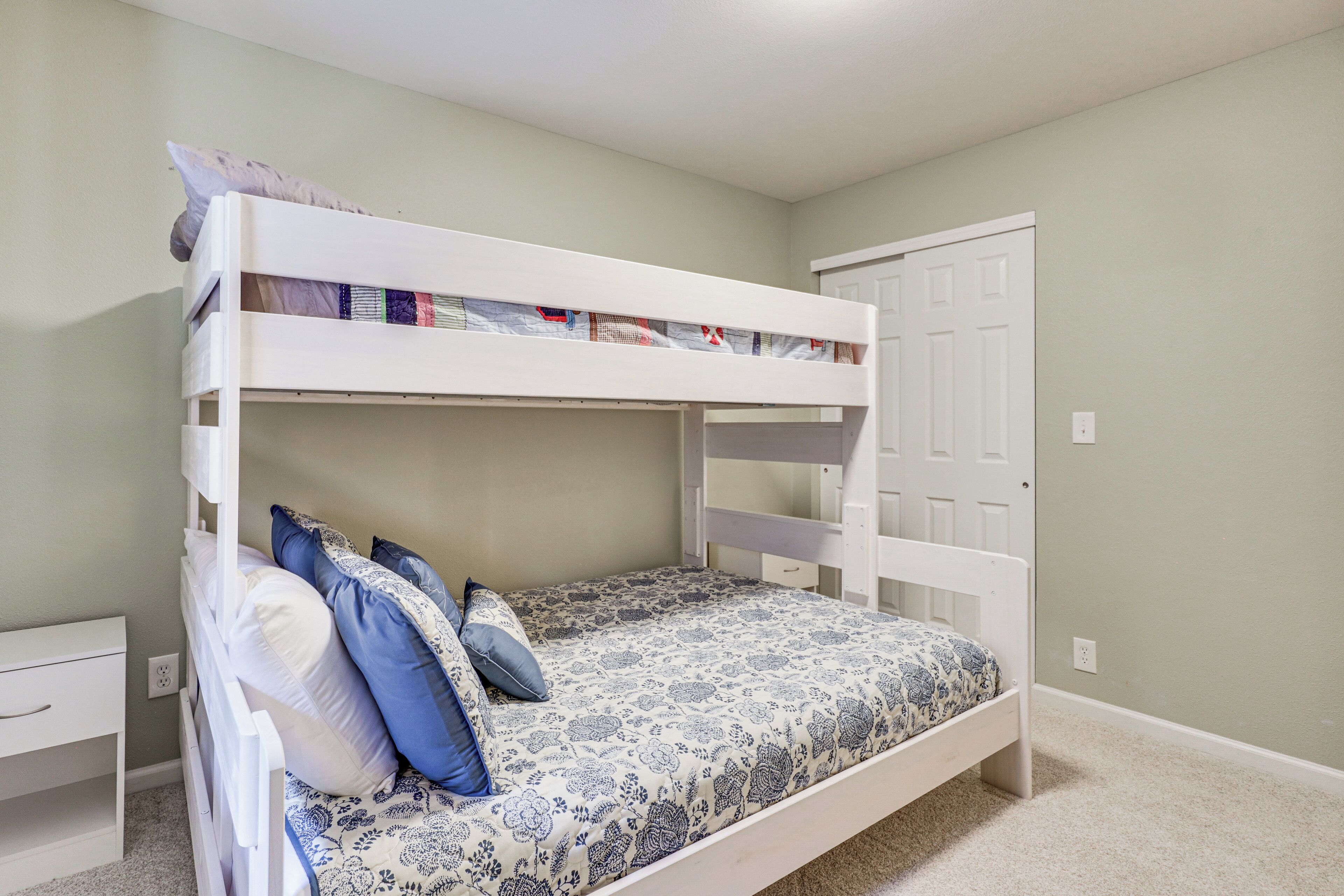 A bunk bed with a patterned bedspread and pillows, a white nightstand, and a door to the right.