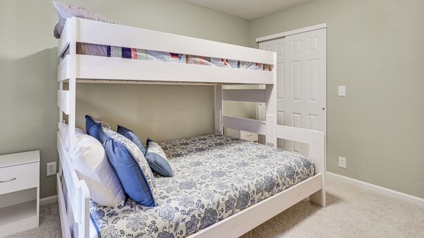 Una cama litera con una colcha y almohadas estampadas, una mesita de noche blanca y una puerta a la derecha.