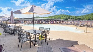 A poolside area with tables and chairs, a covered seating area, and a view of a building across the water.