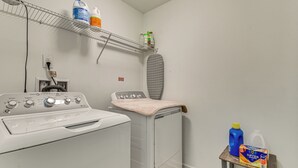 Lavandería con lavadora, secadora, tabla de planchar y estantes con productos de limpieza.