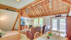 Ein gemütliches Wohnzimmer mit hohen Decken, Holzbalken, einem Kamin, einem Esstisch mit Stühlen und einem Fernseher im Hintergrund.