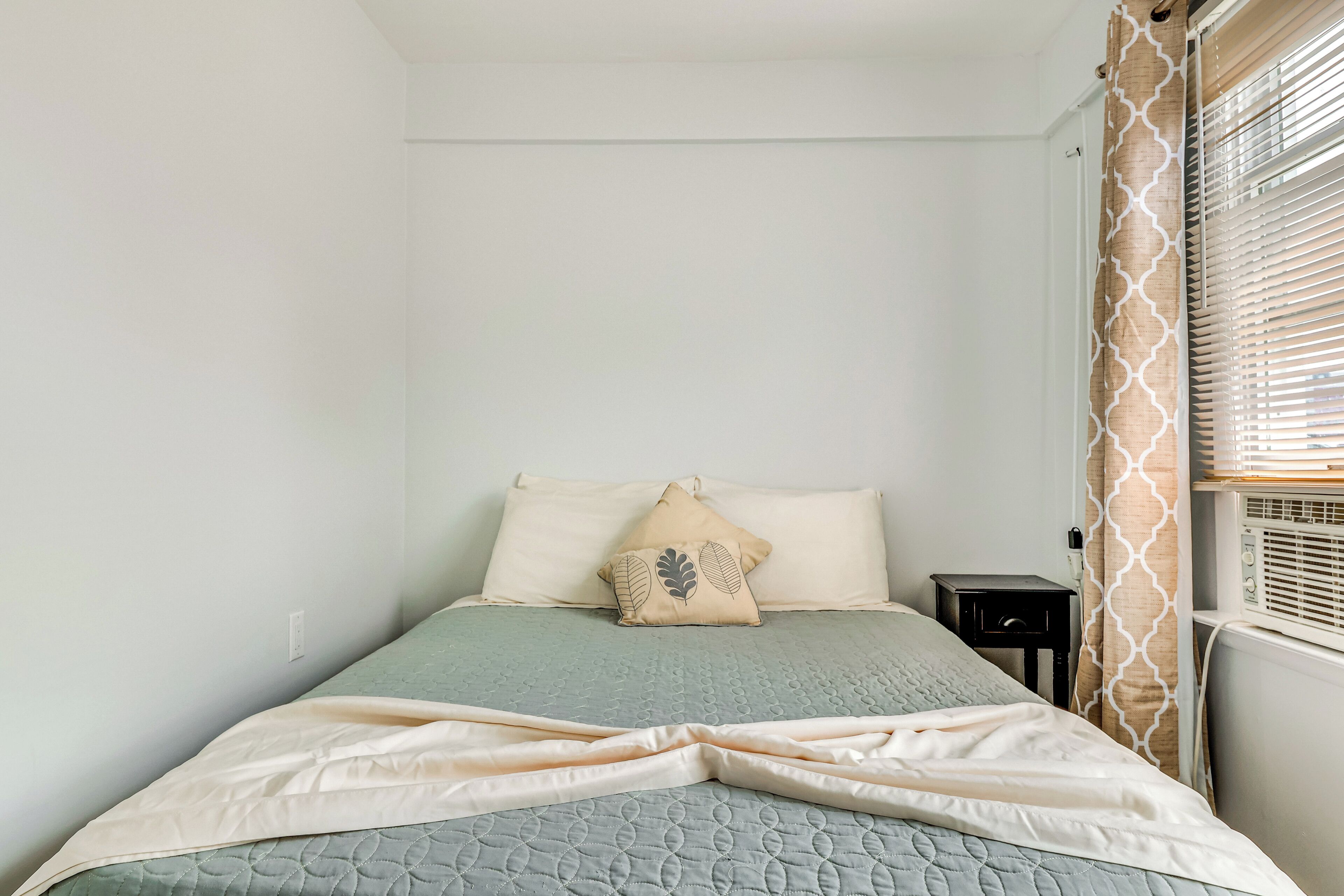 Quarto com cama, almofadas, mesa de cabeceira e janela com persianas.