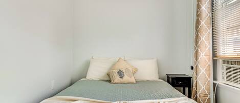 Quarto com cama, almofadas, mesa de cabeceira e janela com persianas.