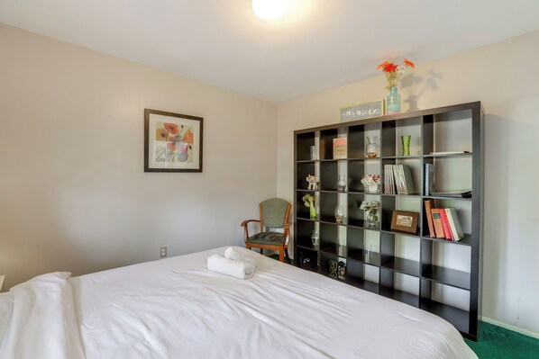 A neatly made bed with white linens, a chair with a cushion, a bookshelf with various items, and a framed picture on the wall.